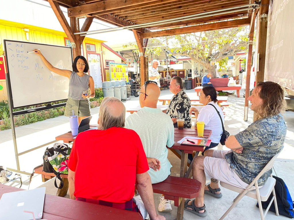 Small-group Thai learning session — instructor at whiteboard teaching tones to seated participants outdoors